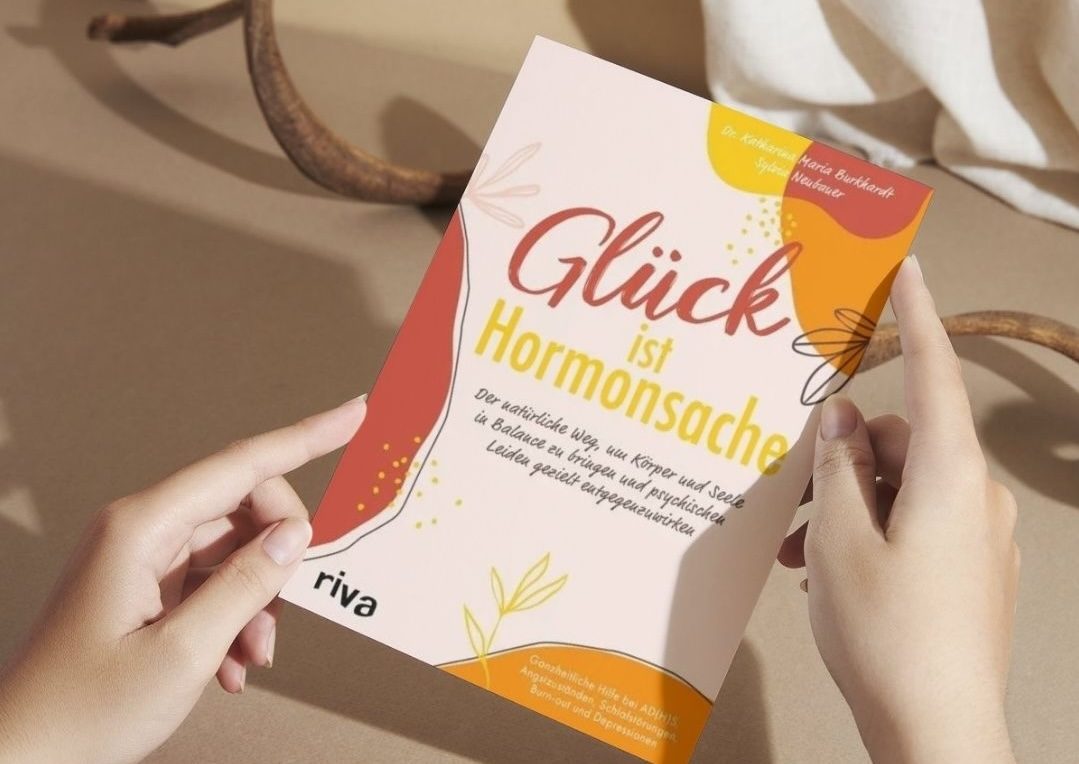 Zwei Hände halten das Buch "Glück ist Hormonsache" vor einem neutralen Hintergrund